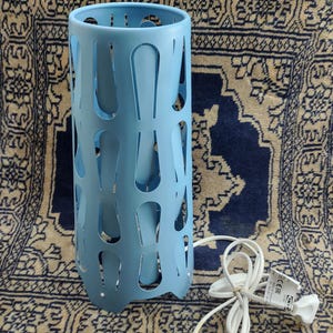 Può includere: Una lampada da tavolo blu chiaro con un design a taglio. La lampada ha un cavo e una spina bianchi con un marchio CE e il testo "IKEA" e "Made in China".