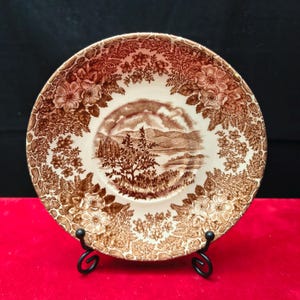 Può includere: Un piatto decorativo marrone e bianco con una scena di paesaggio al centro. Il piatto presenta un bordo floreale ed è esposto su un supporto in metallo nero. Il piatto ha un diametro di circa 20 cm.