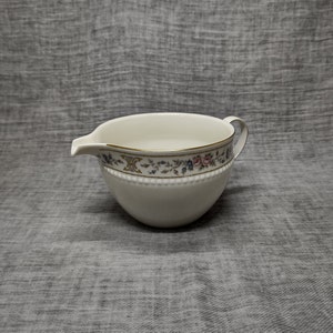 Peut inclure: Un pot à crème en porcelaine blanche avec un motif floral et une bordure dorée. Le pot à crème a une petite poignée et une forme arrondie.