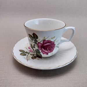 Può includere: Una tazza da tè e un piattino bianchi con un bordo dorato. La tazza e il piattino presentano un motivo floreale con una rosa rosa e foglie verdi.