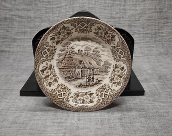 May include: A brown and white ceramic plate with a cottage scene and floral border. The plate is on a black wooden stand.