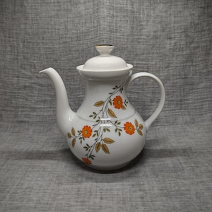 Pode incluir: Bule de cerâmica branca com um design floral com flores laranja e folhas marrons. A chaleira tem uma tampa e uma alça.