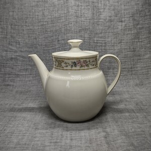 May include: A white porcelain teapot with a lid, featuring a floral and gold band around the top. The teapot has a curved spout and handle, and a gold trim. The background is a gray fabric.