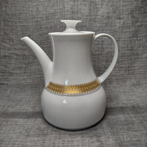 May include: A white ceramic coffee pot with a lid and a handle. The pot features a gold decorative band around the middle. The spout is long and curved. The pot is on a gray background.