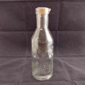 Puede incluir: Botella de vidrio transparente con tapón de corcho. La botella tiene un cuello estrecho y una base más ancha. Se puede ver texto en relieve en la botella. La botella está diseñada para líquidos, probablemente leche o un producto similar.