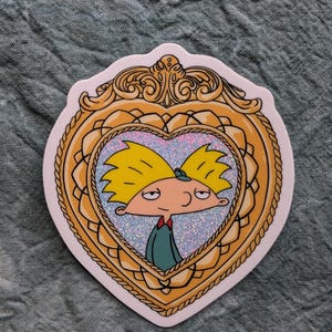 May include: A sticker featuring a cartoon character in a heart-shaped frame. The character has yellow hair, a green shirt, and a red bow tie. The frame is gold with ornate detailing and a glittery background.