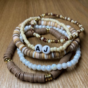 Op de afbeelding: Stapel van vijf handgemaakte armbanden in neutrale tinten. De armbanden hebben een mix van bruine, beige, crème en gouden kralen. Eén armband heeft witte letterkralen met de letters "A J". De armbanden zijn gestapeld op een houten oppervlak.