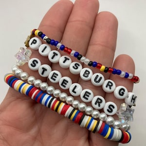 Op de afbeelding: Een set van vijf armbanden met de woorden "Pittsburgh Steelers" in witte kralen, samen met een rood-wit-blauw gestreepte armband en een armband met witte parels.