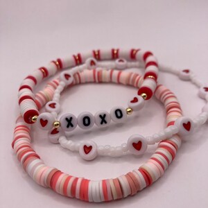 Puede incluir: Conjunto de tres pulseras con cuentas rosas, blancas y rojas. La pulsera del medio tiene cuentas blancas con corazones rojos y letras doradas que deletrean "XOXO".