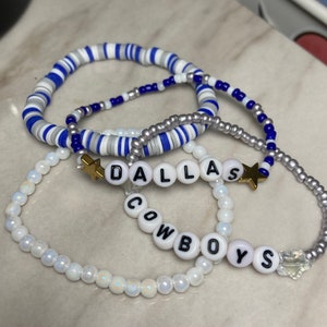 Op de afbeelding: Vier kralenarmbanden met witte en blauwe kralen. De armbanden spellen "Dallas Cowboys" met gouden sterbedels.