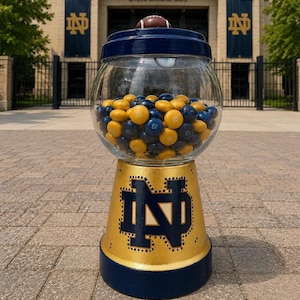 May include: A glass candy dispenser with a navy blue lid topped with a miniature football. The base is painted gold with a navy blue Notre Dame monogram logo and a navy blue bottom rim. The glass globe is filled with navy blue and gold candies.