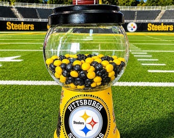 May include: A gumball machine featuring a clear glass globe filled with yellow and black candies. The base is yellow with the Pittsburgh Steelers logo and text. A brown football sits on top of the black lid, set against a football field background.