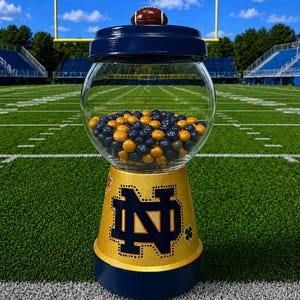 May include: A glass gumball machine with a navy blue lid topped by a miniature football. The base is painted gold with a navy blue Notre Dame monogram logo, a small star, and a shamrock. The clear globe is filled with navy blue and gold candies.