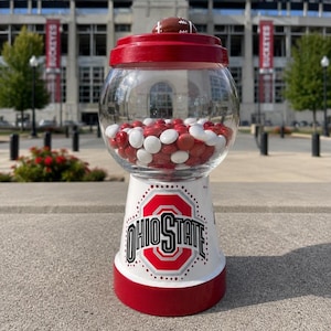 May include: A clear glass gumball machine with a red lid topped by a small brown football. The white base features the Ohio State logo in red, black, and white. The globe is filled with red and white round candies. The design is painted on a pot.