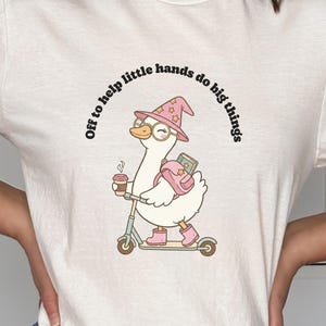 May include: A natural-colored t-shirt featuring a graphic of a white duck wearing a pink star-patterned wizard hat, glasses, and pink boots. The duck carries a pink backpack and rides a scooter while holding a coffee cup. Text reads: Off to help little hands do big things.