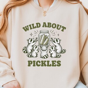 Peut inclure: Un sweat-shirt à col rond couleur sable avec un motif vert foncé représentant quatre ratons laveurs entourant un bocal en verre rempli de cornichons. Le texte au-dessus du bocal indique WILD ABOUT et en dessous PICKLES en police verte.