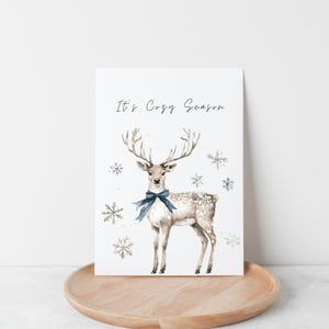May include: A watercolor illustration of a deer with a blue bow tie, surrounded by snowflakes. The text "It's Cozy Season" is written at the top of the image.