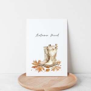 Puede incluir: Una ilustraci&oacute;n de acuarela de un par de botas de goma marrones con hojas de oto&ntilde;o ca&iacute;das. El texto "Autumn Mood" est&aacute; escrito sobre las botas.