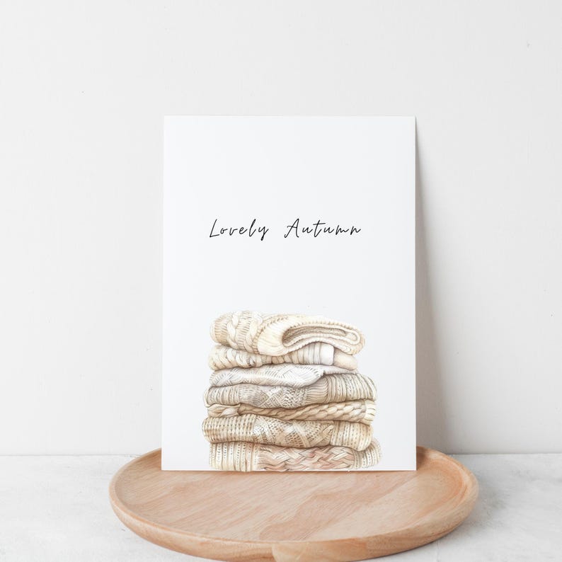 Puede incluir: Una impresi&oacute;n en acuarela con una pila de su&eacute;teres beige doblados. La impresi&oacute;n tiene las palabras "Lovely Autumn" en escritura cursiva. La impresi&oacute;n est&aacute; sobre un fondo blanco y sobre una bandeja de madera.