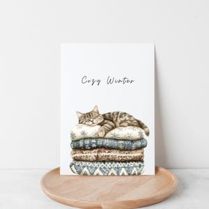 May include: A watercolor illustration of a cat sleeping on a stack of cozy blankets with the text "Cozy Winter" above the cat.