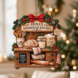 May include: A decorative hanging ornament shaped like a kitchen counter with a loaf of bread, a flour sack, a rolling pin, and kitchen tools. The sign reads ANNA'S SOURDOUGH KITCHEN, 2026, and Baked with Love. It features a red bow and pine greenery.