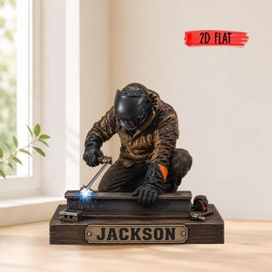 May include: A figurine of a welder wearing a brown jacket, black gloves, and a black helmet, kneeling while welding a metal beam. The base is rectangular with a metal plaque reading JACKSON. A red graphic in the corner reads 2D FLAT.