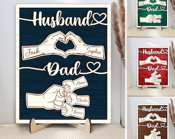 May include: A rectangular wooden sign featuring laser-cut designs on a dark blue wood-grain background. The text "Husband" and "Dad" are displayed with heart icons. Two hands form a heart shape with names "Jack" and "Sophia," and a fist bump graphic includes smaller hands with names "Anna," "Charlie," and "Becca."