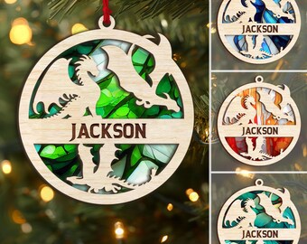 May include: Wooden Christmas ornament with a dragon design and the name "JACKSON" in brown lettering. The ornament has a circular shape with a red ribbon for hanging. The background features a stained glass effect with green and white colors.