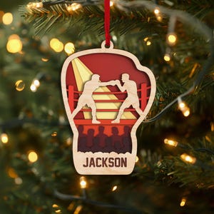 May include: Wooden Christmas ornament shaped like a boxing glove, featuring a boxing ring scene with two boxers. The ornament has a red ribbon for hanging and the name "JACKSON" at the bottom. The background is a blurred image of a Christmas tree with lights.