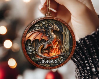May include: A round, wooden Christmas ornament featuring a detailed dragon design. The dragon is depicted with orange wings and is standing in front of a fire. The name "JACKSON" is displayed at the bottom. The ornament is held by a gold string.