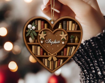 May include: A heart-shaped wooden ornament featuring a bookshelf design. The ornament has a smaller heart in the center with the name "Sophia" and decorative elements. The shelves are filled with miniature books, plants, and other decorative items. A gold string is attached for hanging.