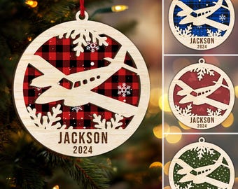 Könnte beinhalten: Weihnachtsbaumschmuck aus Holz mit Flugzeugdesign, Schneeflocken und dem Namen "JACKSON" und dem Jahr "2024". Der Schmuck hat einen rot-schwarzen Karohintergrund. Ein rotes Band ist zum Aufhängen befestigt.