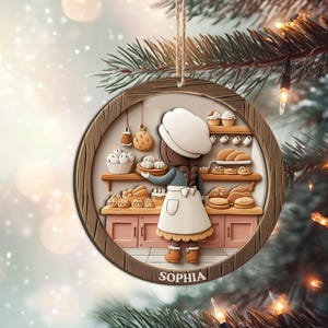 May include: A round wooden Christmas ornament featuring a baker in a bakery. The baker is wearing a white apron and hat, holding a tray of pastries. The ornament has a brown wooden frame and the name "SOPHIA" at the bottom.