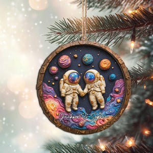 May include: A round Christmas ornament featuring two astronauts holding hands in a space-themed design. The ornament has a wooden frame and a string for hanging. The names "Jackson & Sophia" are written on the bottom.