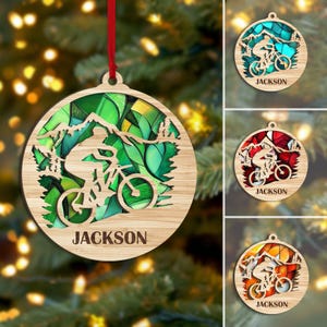 May include: Wooden Christmas ornament featuring a mountain biker design with the name "JACKSON" at the bottom. The ornament has a stained glass effect with green, yellow, and blue hues. A red ribbon is attached for hanging.