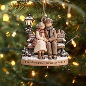 May include: A hanging Christmas ornament featuring a couple sitting on a bench under a streetlamp. The woman is wearing a cream coat and red scarf, the man a brown coat and grey scarf. The base reads, From Our First Kiss Till Our Last Breath.
