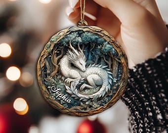 May include: A round, wooden Christmas ornament featuring a white dragon nestled in a forest scene. The dragon has a detailed, scaled body and is surrounded by trees and foliage. The name "Sophia" is inscribed on the ornament.