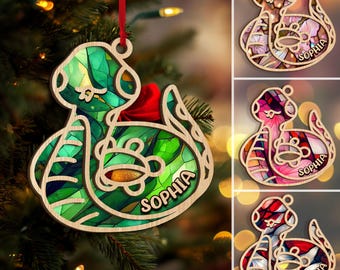 Può includere: Ornamento natalizio in legno a forma di serpente con un design a vetrata. L'ornamento è verde, rosso e marrone, con il nome "Sophia" stampato sopra. È appeso a un albero di Natale.