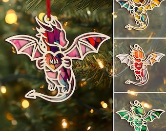 May include: Wooden dragon ornament with a stained glass design. The dragon has outstretched wings and a heart-shaped body with the name "MIA". The ornament hangs from a red ribbon and is displayed on a Christmas tree.