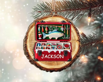 May include: A red fishing tackle box Christmas ornament with the name "JACKSON" on the front. The ornament is shaped like a wooden slice and features a detailed interior with fishing lures, a fish, and spools of thread. The ornament hangs from a twine string.