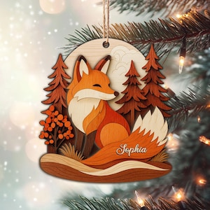 May include: Wooden Christmas ornament featuring a layered design of a fox in a forest scene. The fox is orange and white, with a fluffy tail. The name "Sophia" is written on the tail. The background includes trees, a moon, and a snowy effect.