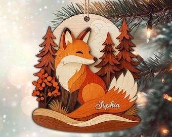 May include: Wooden Christmas ornament featuring a layered design of a fox in a forest scene. The fox is orange and white, with a fluffy tail. The name "Sophia" is written on the tail. The background includes trees, a moon, and a snowy effect.