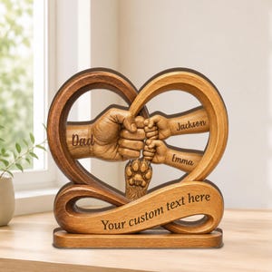 May include: Wooden heart-shaped family sculpture with the names "Dad", "Jackson", and "Emma" carved into it. The sculpture features hands clasped together, with a paw print and the text "Your custom text here" on the base.