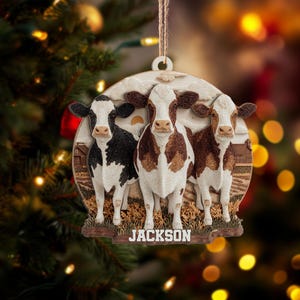 May include: A round Christmas ornament featuring three cows in a farm scene. The cows are black and white, brown and white, and brown. The word "JACKSON" is at the bottom. The ornament hangs from a twine string.