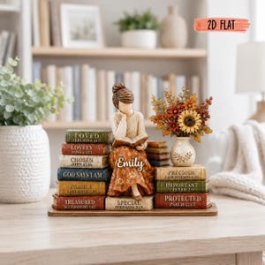 May include: A figurine of a person reading a book while sitting on a stack of books labeled with words like Loved, Lovely, Chosen, God Says I Am, Family, Treasured, Emily, Special, Precious, Important, and Protected, with a small flower vase.