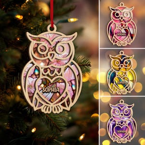 May include: Wooden owl ornament with a floral design and the name "Sophia" in a heart-shaped cutout. The ornament hangs from a red ribbon. Additional owl ornaments in pink, yellow, and purple are also shown.