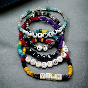 May include: A set of six colorful beaded bracelets with different words spelled out on them. The words are "Promise", "Flie", "Filter", "Serendip", and "Vibe".