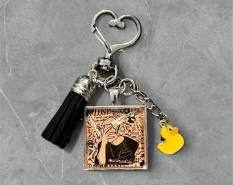 May include: A silver-toned keychain with a heart-shaped clasp, a black tassel, and a square pendant featuring a graphic of a person covering their face. A small yellow rubber duck charm hangs from the chain.