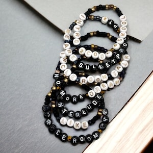 Może przedstawiać: Zestaw 10 bransoletek z czarnymi i złotymi koralikami. Każda bransoletka ma inne słowo napisane białymi koralikami, w tym "Intro", "Wear", "Circle", "No More", "Interlude", "Like", "2 Cool", "Bullet", "Dream" i "Talk".