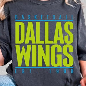 May include: A charcoal grey short-sleeve t-shirt featuring the text "BASKETBALL" in light blue, "DALLAS WINGS" in large lime green letters, and "EST. 1998" in light blue. The shirt has a relaxed fit and a vintage-washed appearance.